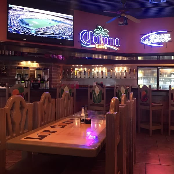 Interior of Los Cocos Mexican Restaurant with a large TV, neon signs, bar, and wooden tables.
