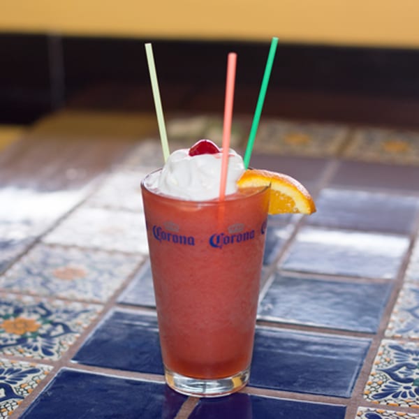 A red frozen drink with whipped cream, a cherry, and an orange slice in a Corona glass.