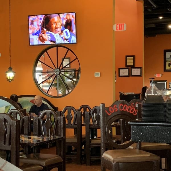Interior of Los Cocos Mexican Restaurant with orange walls, a TV, wagon wheel decor, and diners.