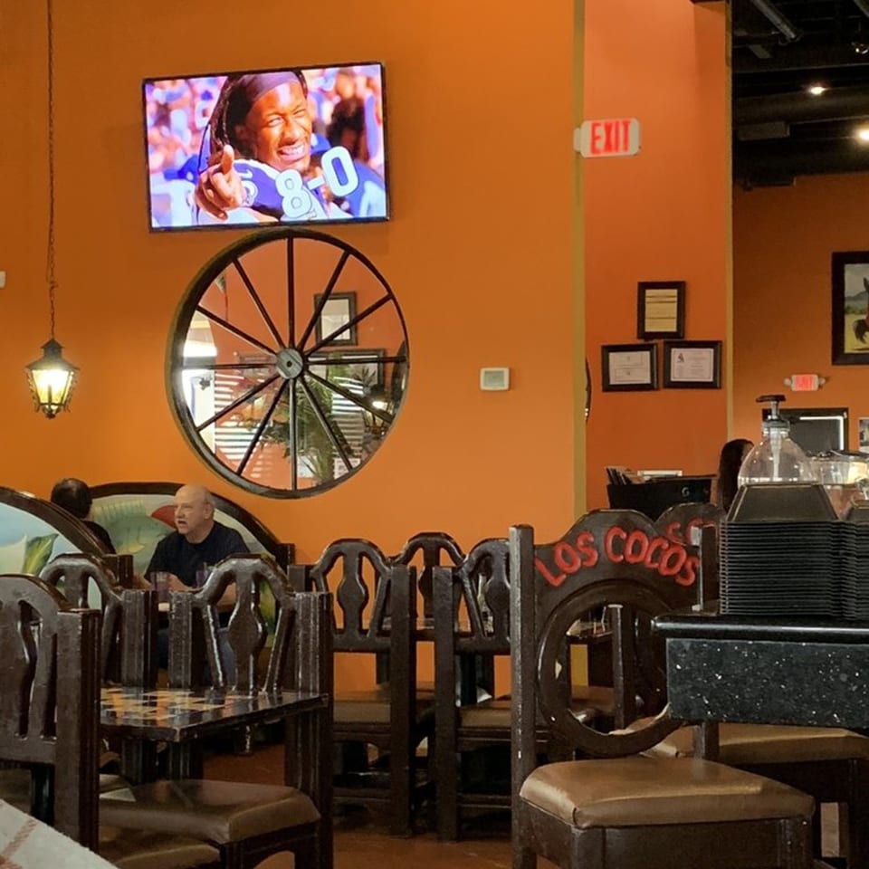 Interior of Los Cocos Mexican Restaurant with orange walls, a TV, wagon wheel decor, and diners.