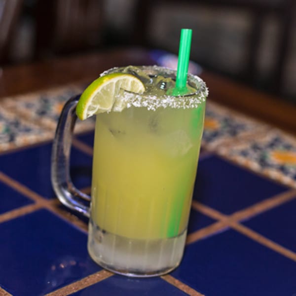 A salted margarita with a lime wedge and green straw in a glass mug on a blue tiled table.