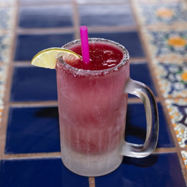 A frosty mug of red frozen margarita with a lime wedge and pink straw on a tiled table.