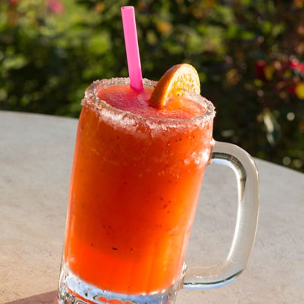 A vibrant red frozen drink in a mug with a salted rim, orange slice, and pink straw.