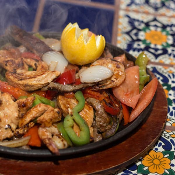 Sizzling fajitas with chicken, shrimp, steak, peppers, onions, and a lemon on a decorative tile table.
