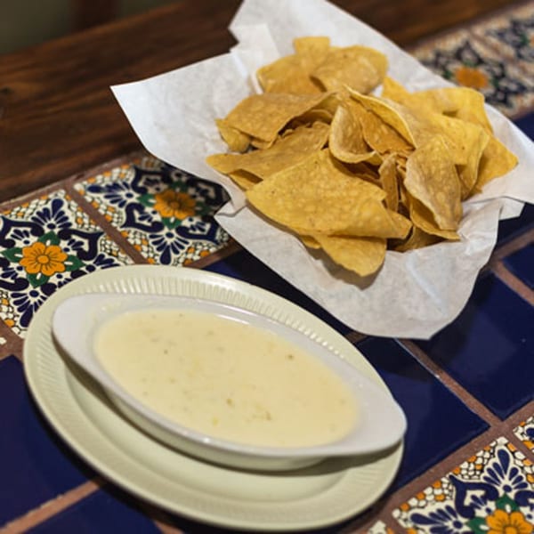A basket of tortilla chips and a bowl of queso dip on a colorful tiled table.