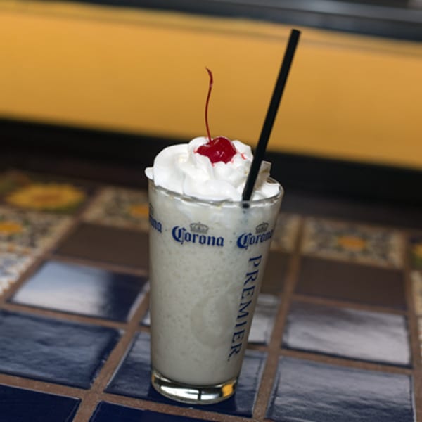 A creamy white drink with whipped cream and a cherry on top, served in a Corona glass.