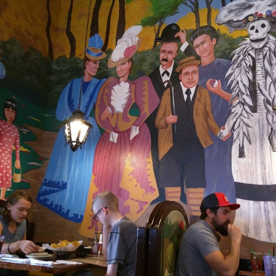 A vibrant mural of people in traditional Mexican attire and a skeleton adorns the restaurant wall.