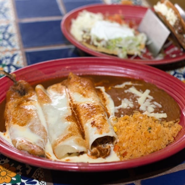 A red plate of Los Cocos Mexican Restaurant's enchiladas, refried beans, and rice, with another dish blurred in the background.