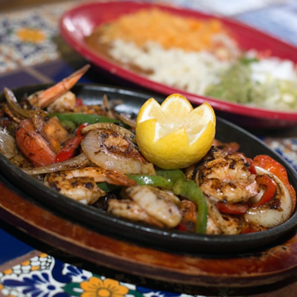 Sizzling shrimp fajitas with bell peppers, onions, and a lemon wedge, with a blurred dish in the background.