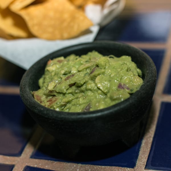 A black bowl of chunky guacamole with red onion sits on a blue tiled table, with tortilla chips in the background.